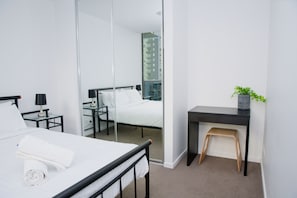 Quarto de hotel moderno com cama, pequena escrivaninha, cadeira, espelho e planta em vaso.