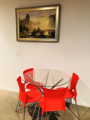 Uma área de refeições com mesa de vidro redonda e cadeiras vermelhas, um quadro de paisagem emoldurado na parede e um interruptor de luz na parede.