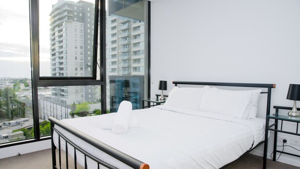 Quarto de hotel moderno com uma janela grande, uma cama bem arrumada e uma mesinha de cabeceira com uma luminária.