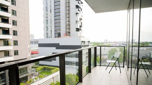 Uma varanda moderna com guarda-corpo de vidro, cadeiras brancas e vista para edifícios e áreas verdes ao redor.