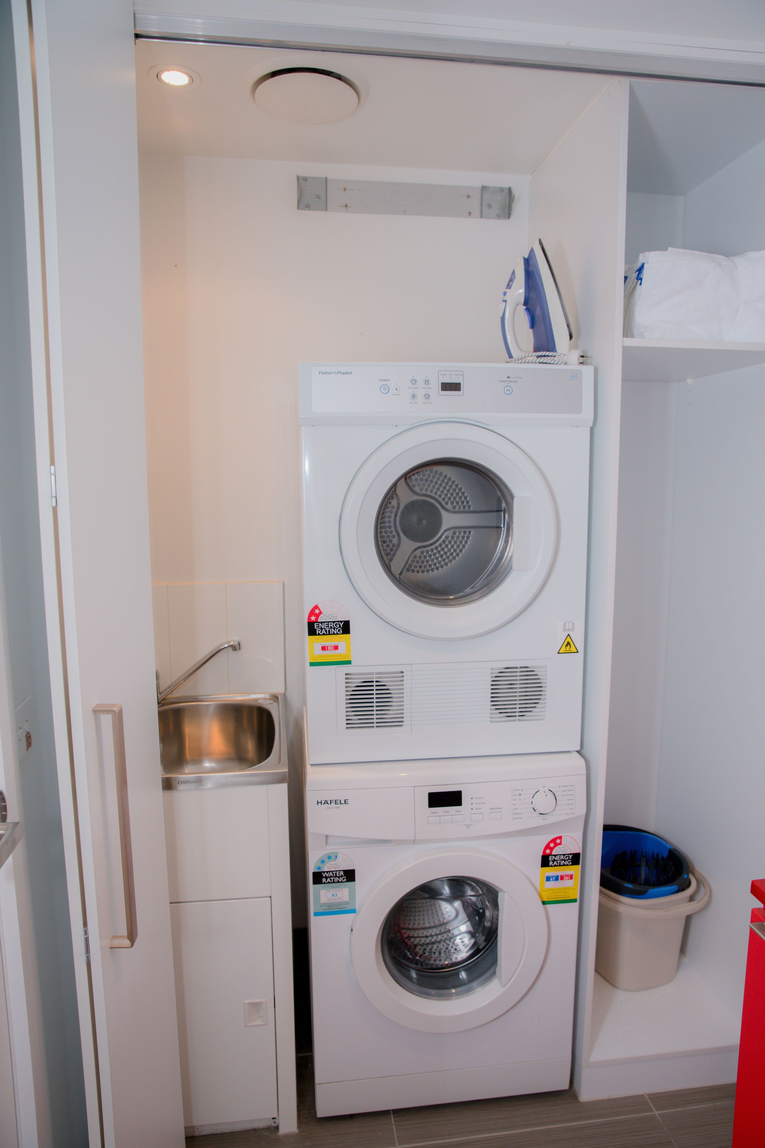 A compact laundry area with a stacked washer and dryer, a small sink, and a trash bin.