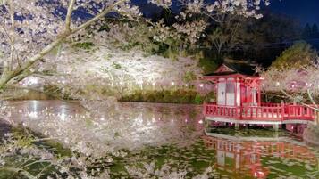 A traditional Japanese-style building with a red roof and white walls, surrounded by a pond with lotus flowers and cherry blossoms.