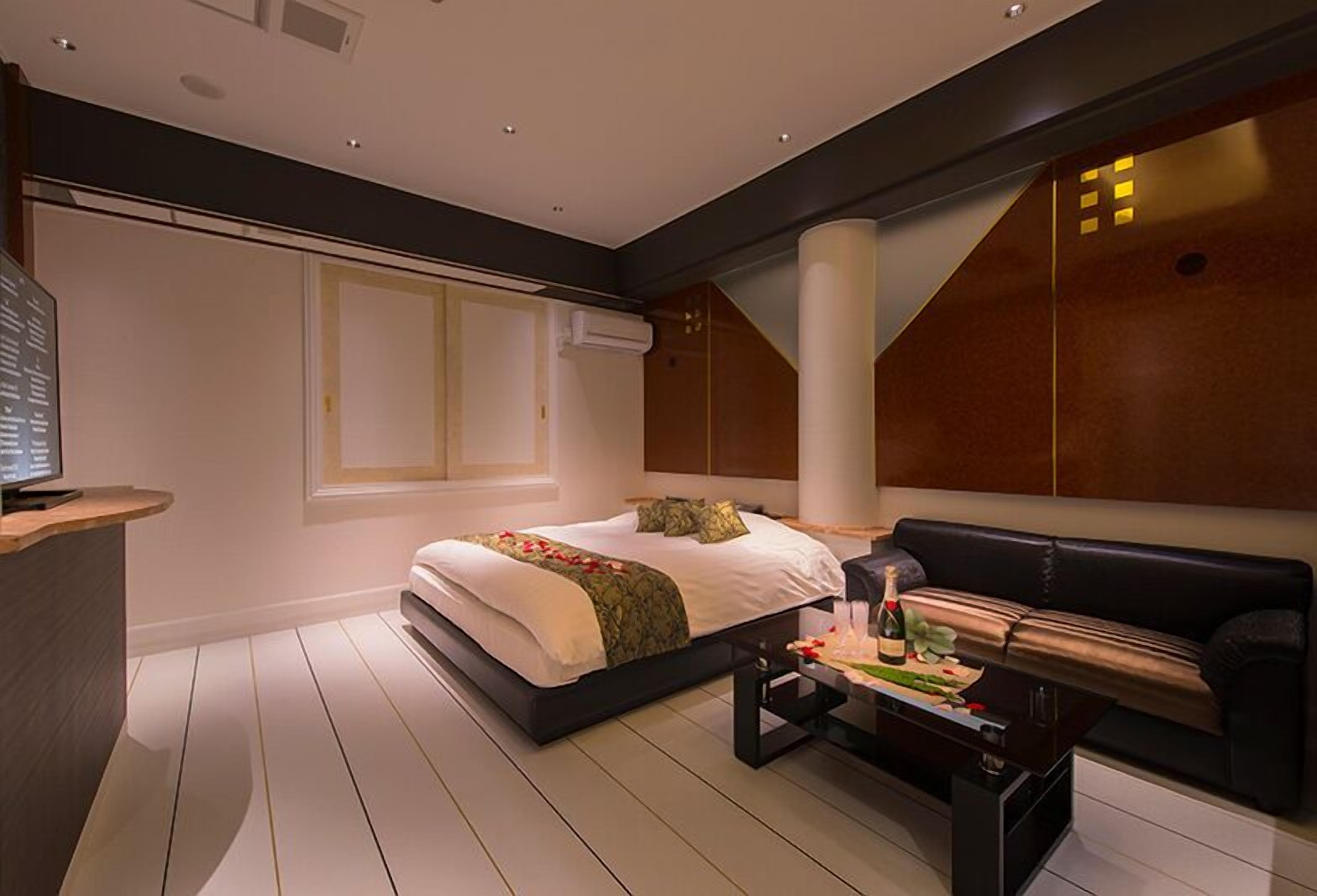 Quarto moderno com uma cama grande, um sofá de couro preto, uma mesa de centro de madeira e uma TV sobre um suporte.