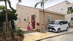 A white SUV parked in front of a brick building with a red roof, a wooden door, and a satellite dish on the roof. - Ecot Shimozato 1 (Miyakojima)
