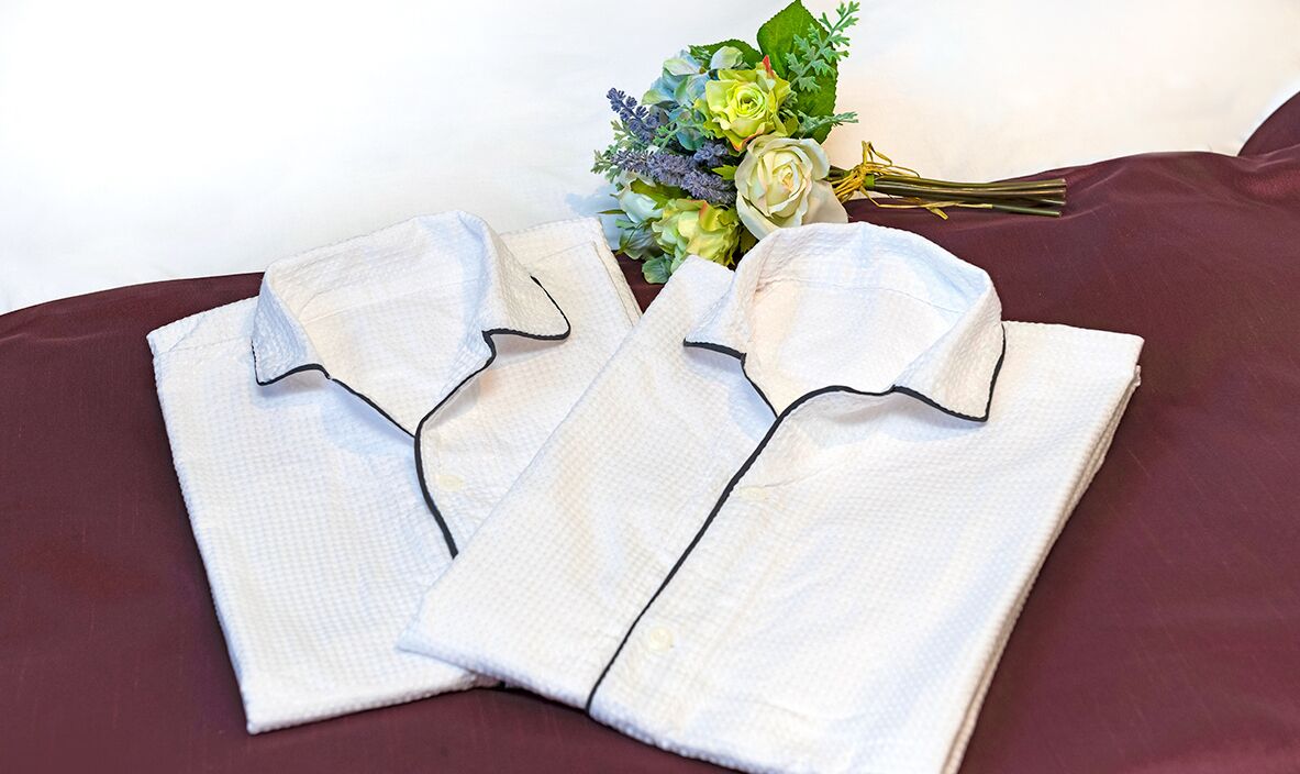 Two neatly folded white shirts with black piping on a dark red surface, accompanied by a bouquet of flowers.