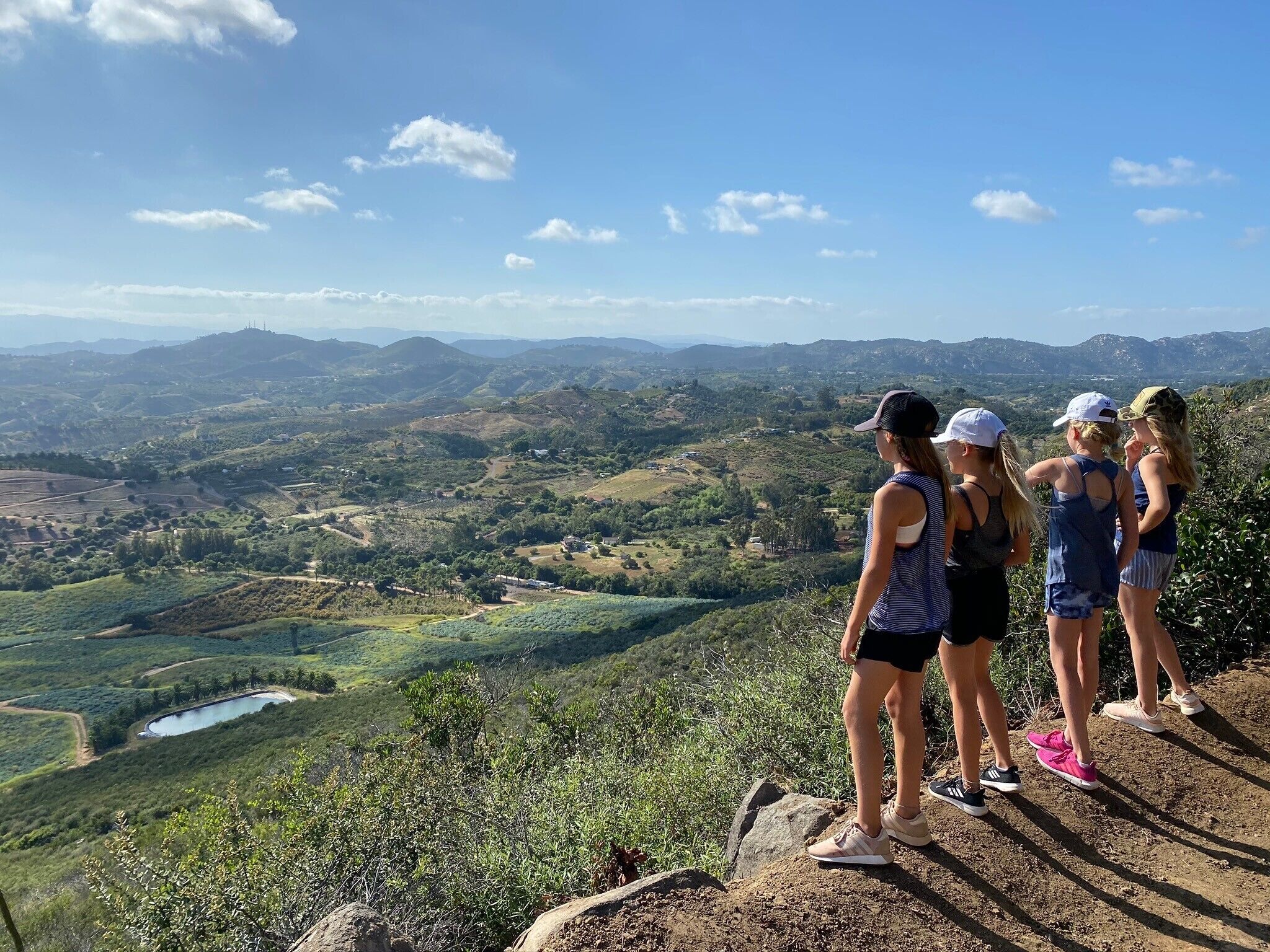 A group of people standing on a rocky outcrop overlooking a valley with a lake and distant hills.