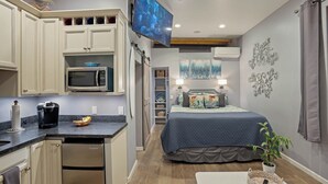 Quarto de hotel compacto com cozinha compacta, uma cama com colcha azul, uma planta em vaso e obras de arte nas paredes.