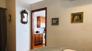 A bedroom with a bed, a green blanket, a wooden door leading to a kitchen, and two framed pictures on the wall.