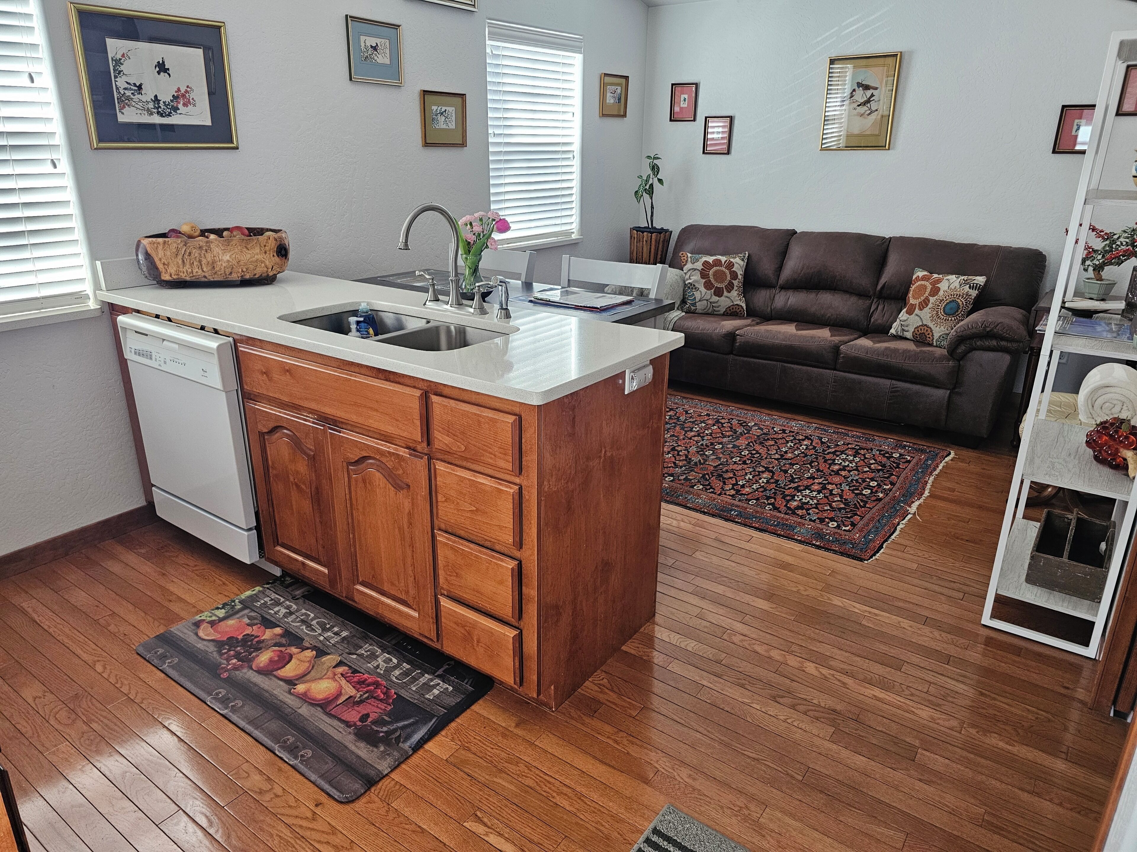 A kitchen with a dishwasher, wooden cabinets, a sink, and a countertop. There is a brown sofa and a patterned rug in the living area.