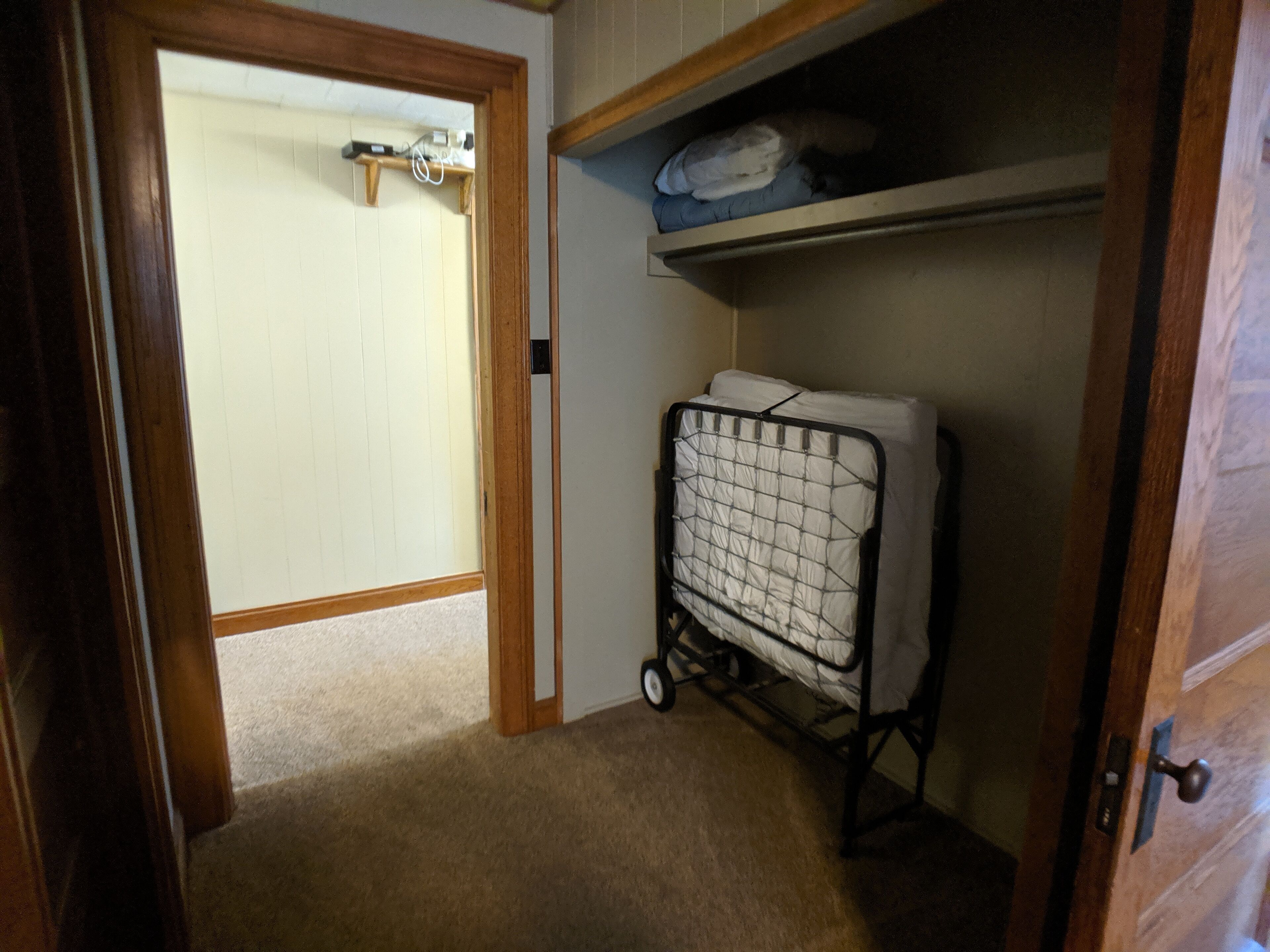 A room with a wooden door, a white wall, a carpeted floor, a metal cart with a mattress, and a shelf with folded blankets.
