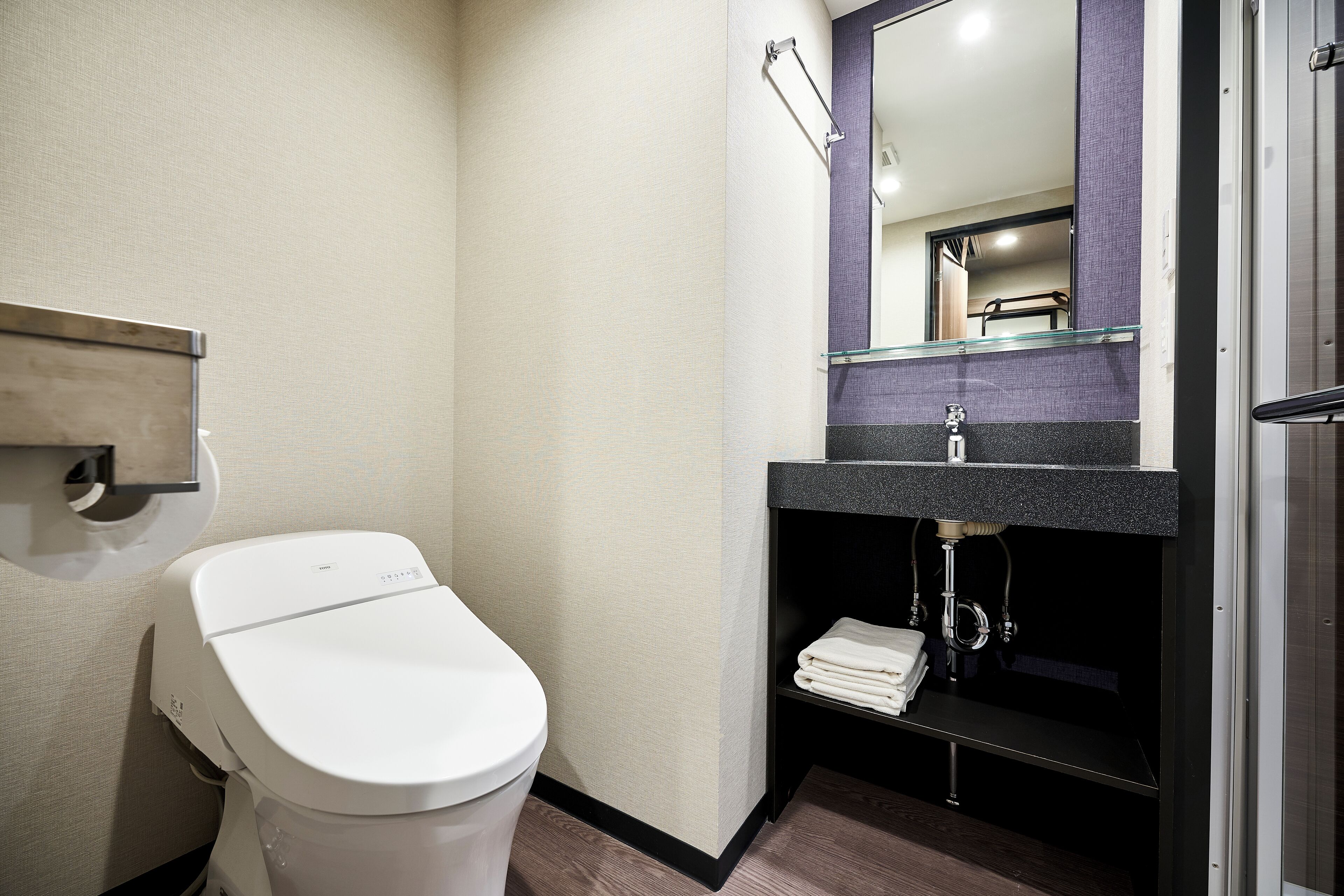 A modern bathroom with a white toilet, a glass shelf with a towel, and a mirror above the sink.