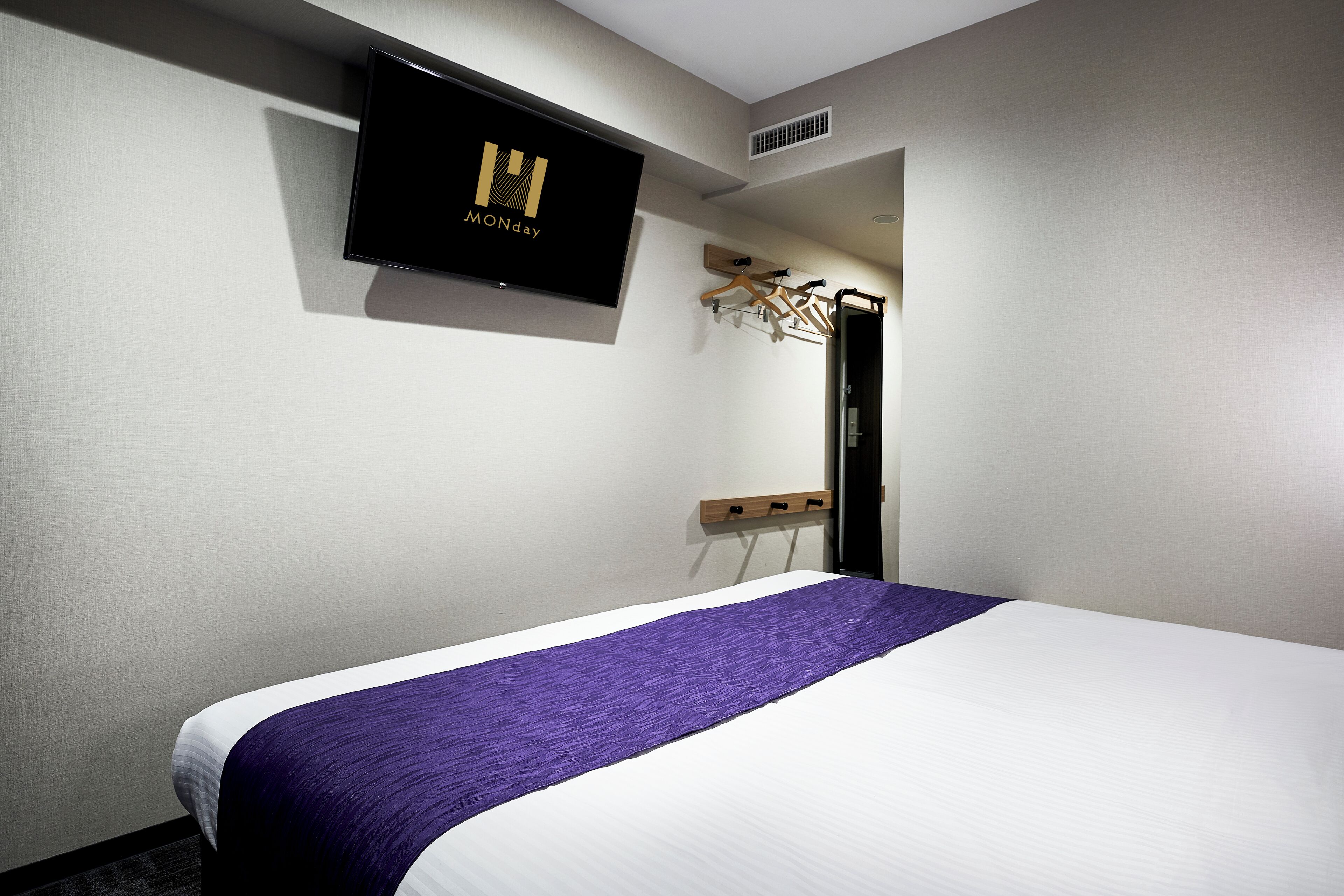 A hotel room with a bed, a TV mounted on the wall, and a coat rack.