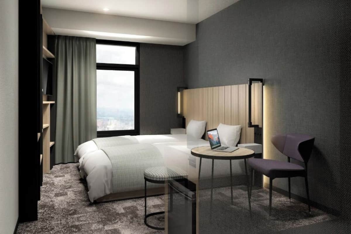 A hotel room with a bed, a desk, a chair, a window, and a view of the city.