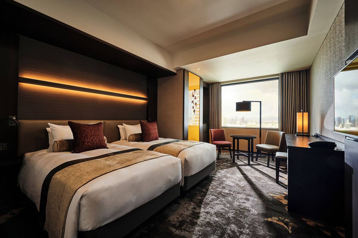 A hotel room with two beds, a desk, a chair, a large window with a city view, and a flat-screen TV.
