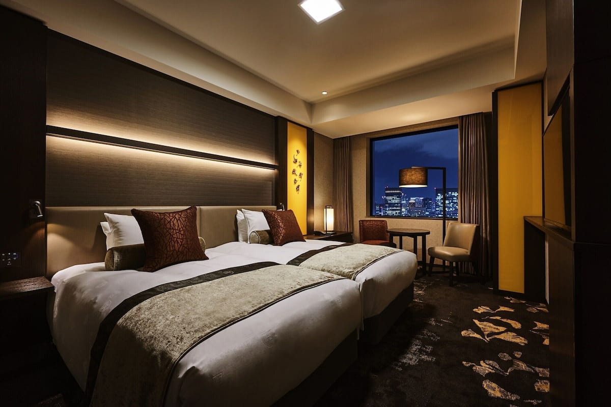 A hotel room with two beds, a nightstand, a chair, a small table, and a view of a cityscape at night.