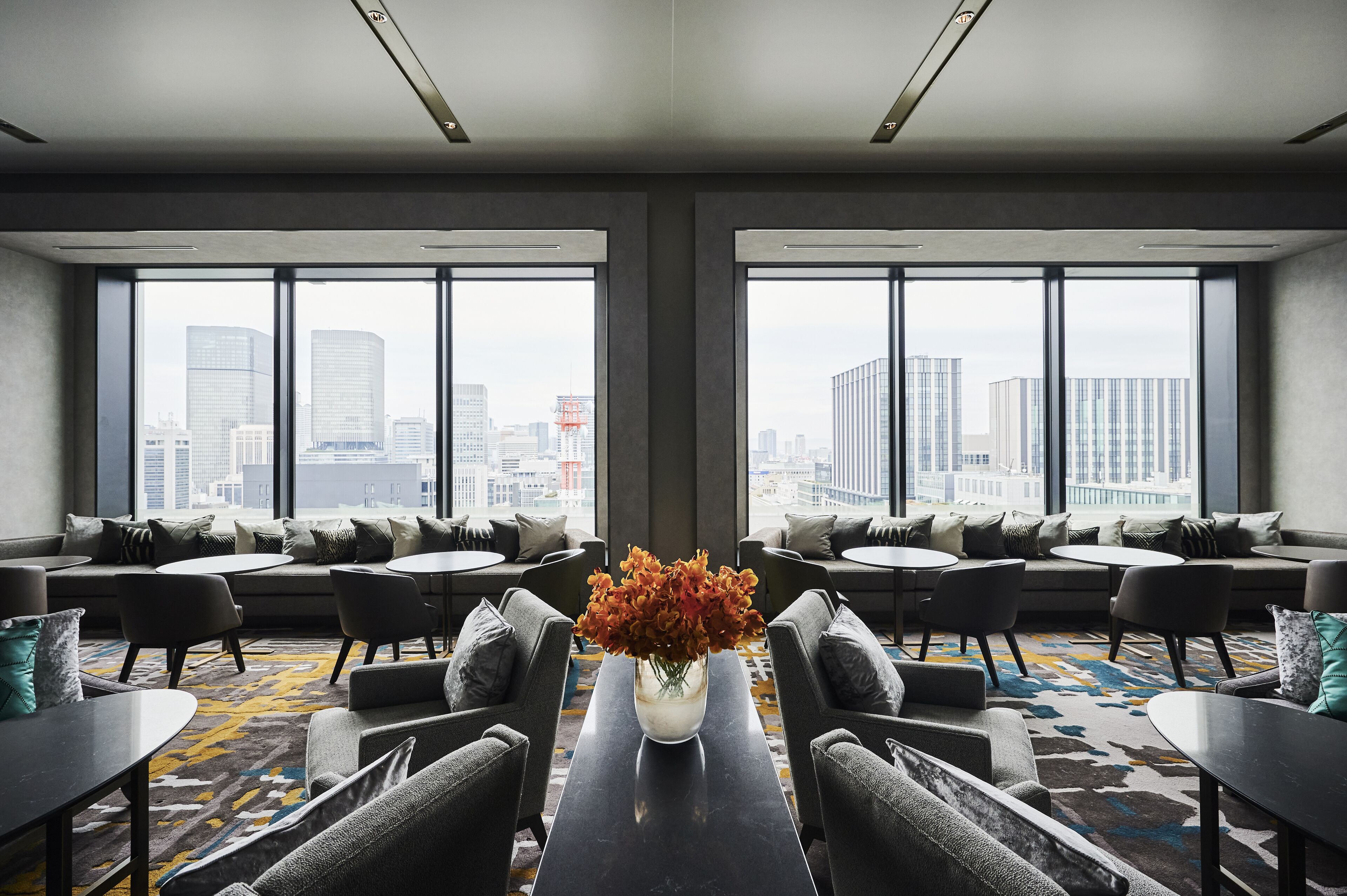 A modern hotel lobby with a large window offering a city view, a central seating area with tables, and a decorative vase with flowers.