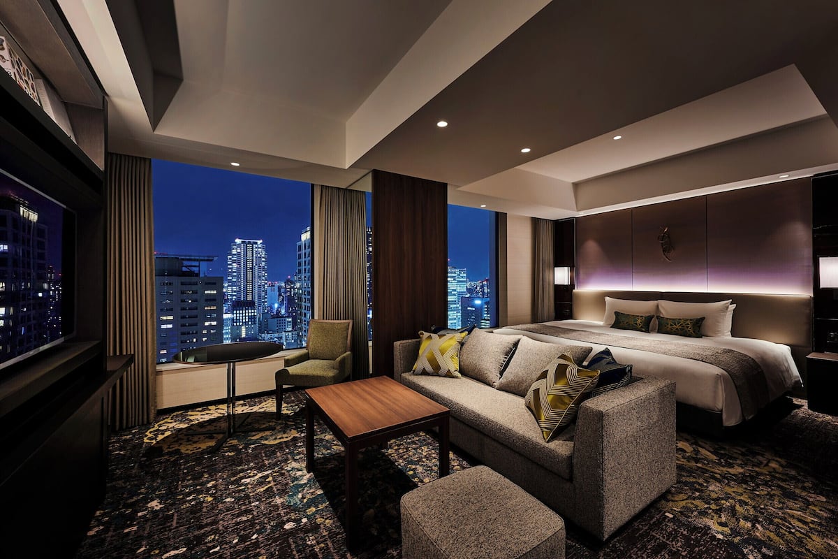 A modern hotel room with a large bed, a sofa, a coffee table, and a TV. The room has a city view and a patterned carpet.