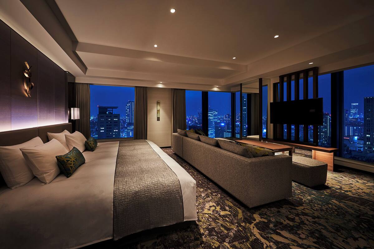 A modern hotel room with a large bed, a sofa, a TV, and a city view.