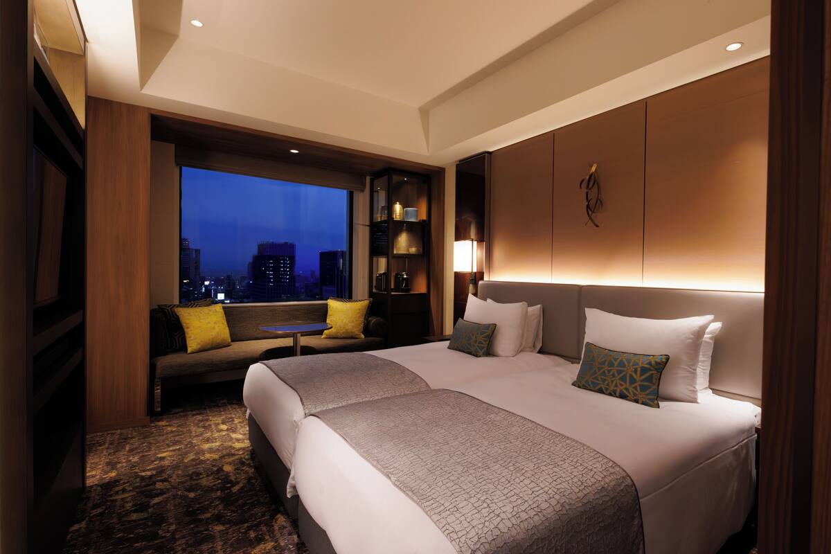 A modern hotel room with two beds, a large window with a city view, a seating area with a table, and a bookshelf.