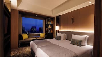 A modern hotel room with two beds, a large window with a city view, a seating area with a table, and a bookshelf.