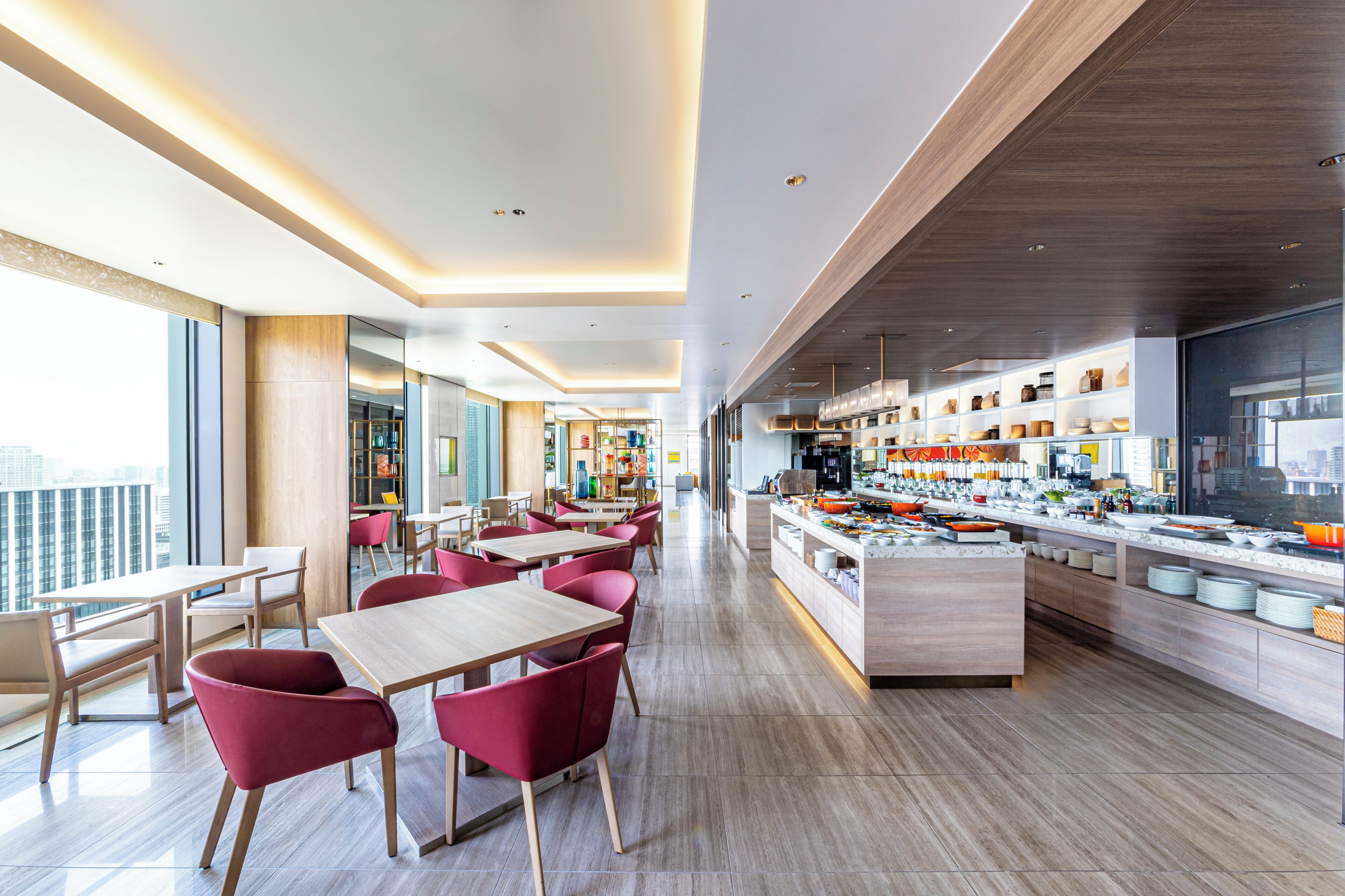 Un espace repas moderne avec des tables en bois et des chaises rouges, une cuisine équipée de différents ustensiles et de grandes fenêtres offrant une vue sur la ville.