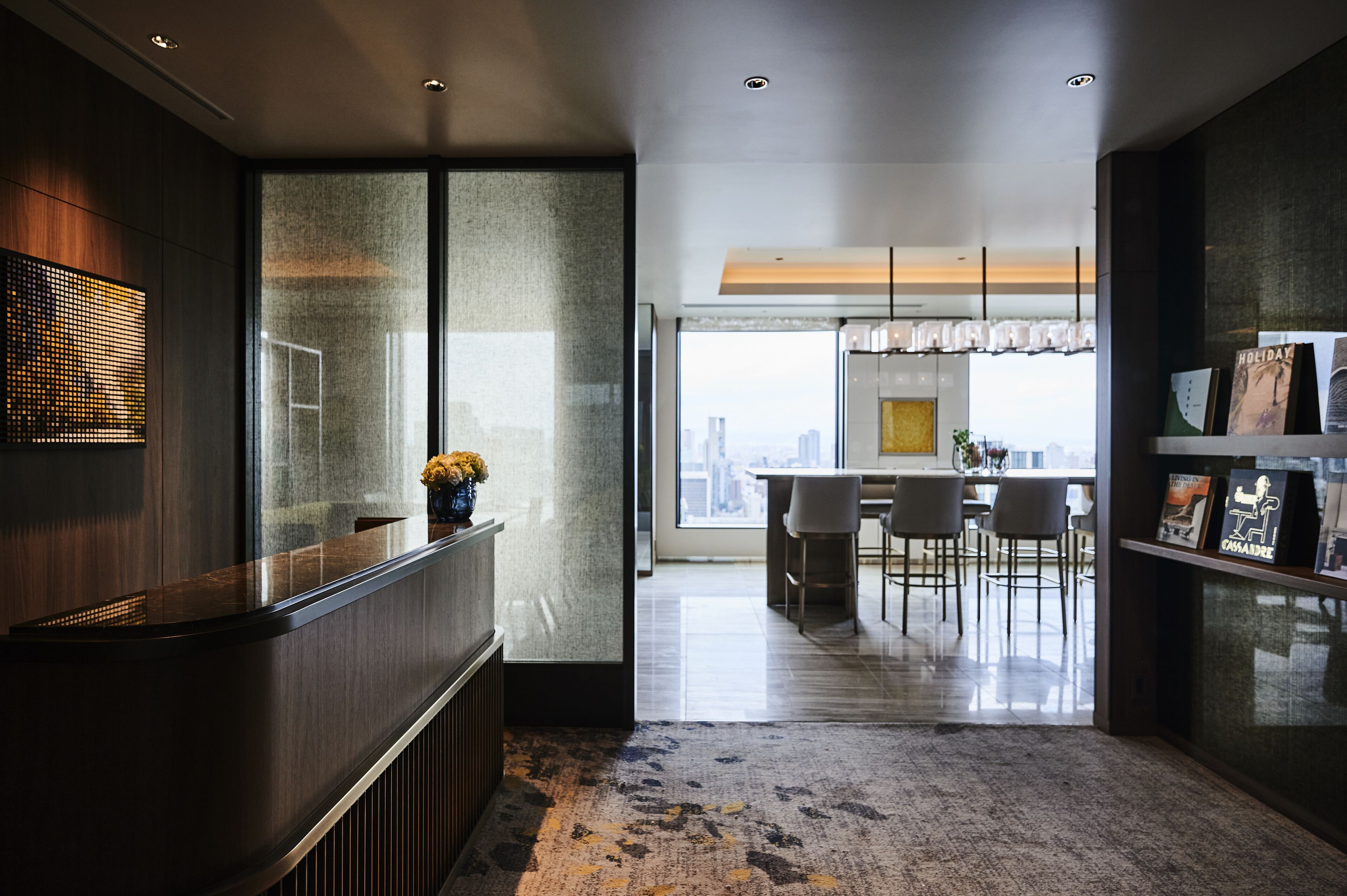 A modern reception area with a glass partition, a reception desk, and a view of a dining area with chairs and a table.