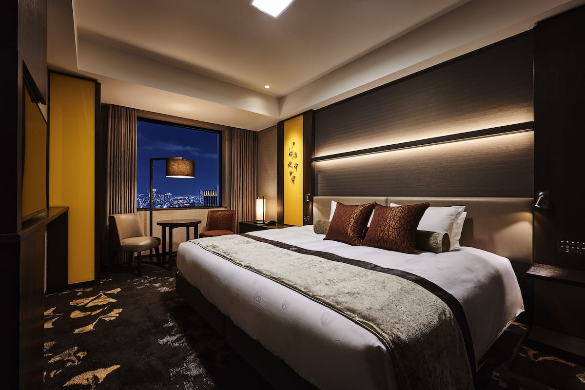 A hotel room with a large bed, a desk, a chair, a nightstand, and a view of the city at night.