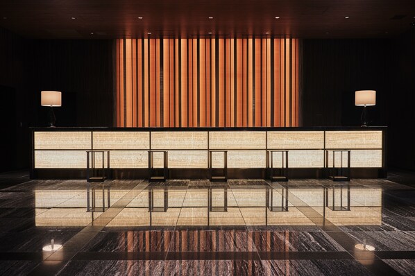 Zona de recepción con piso de mármol pulido, un mostrador de recepción con un panel de luz vertical y dos lámparas a cada lado.