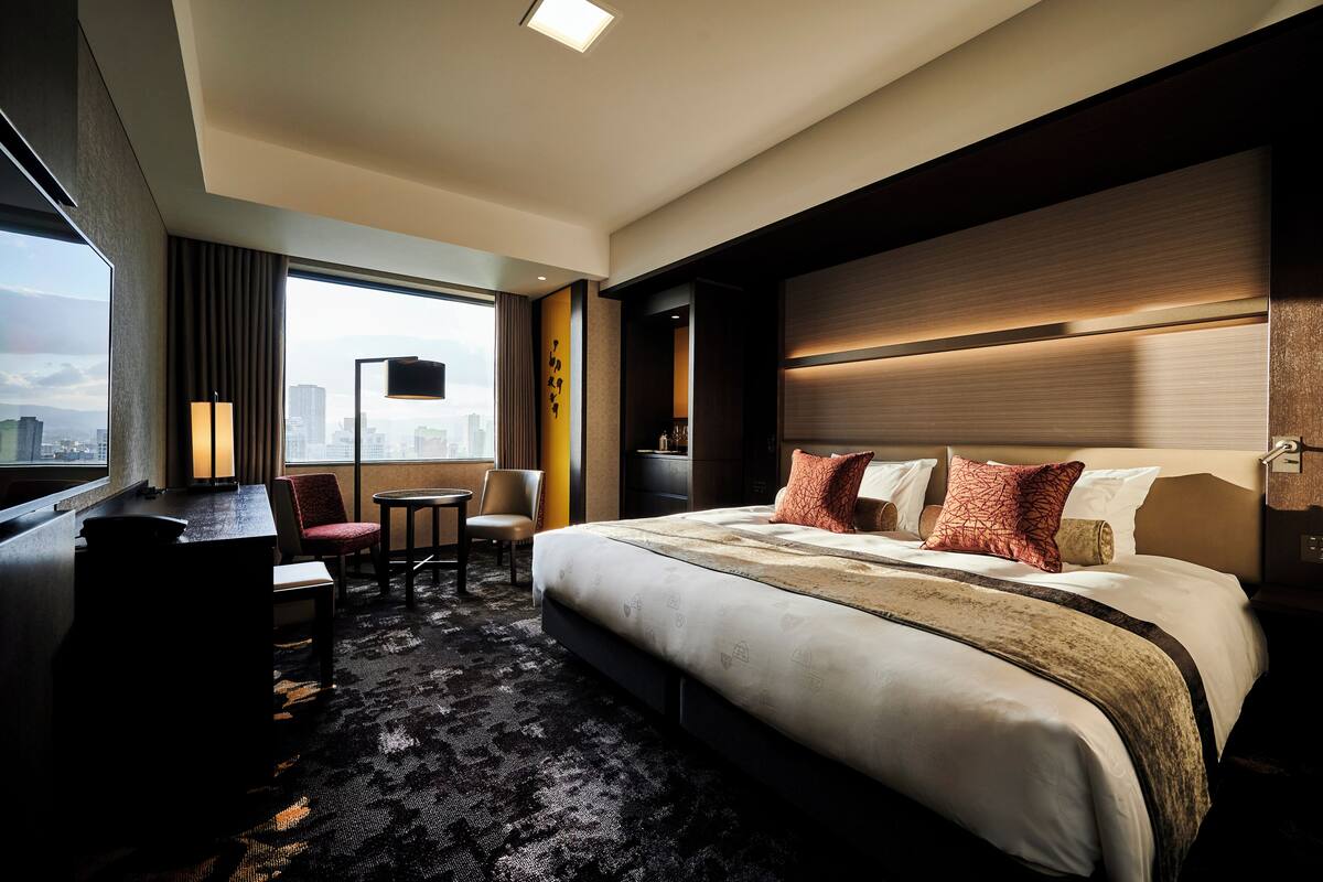 A hotel room with a large bed, a desk, and a chair. There is a city view through the window.