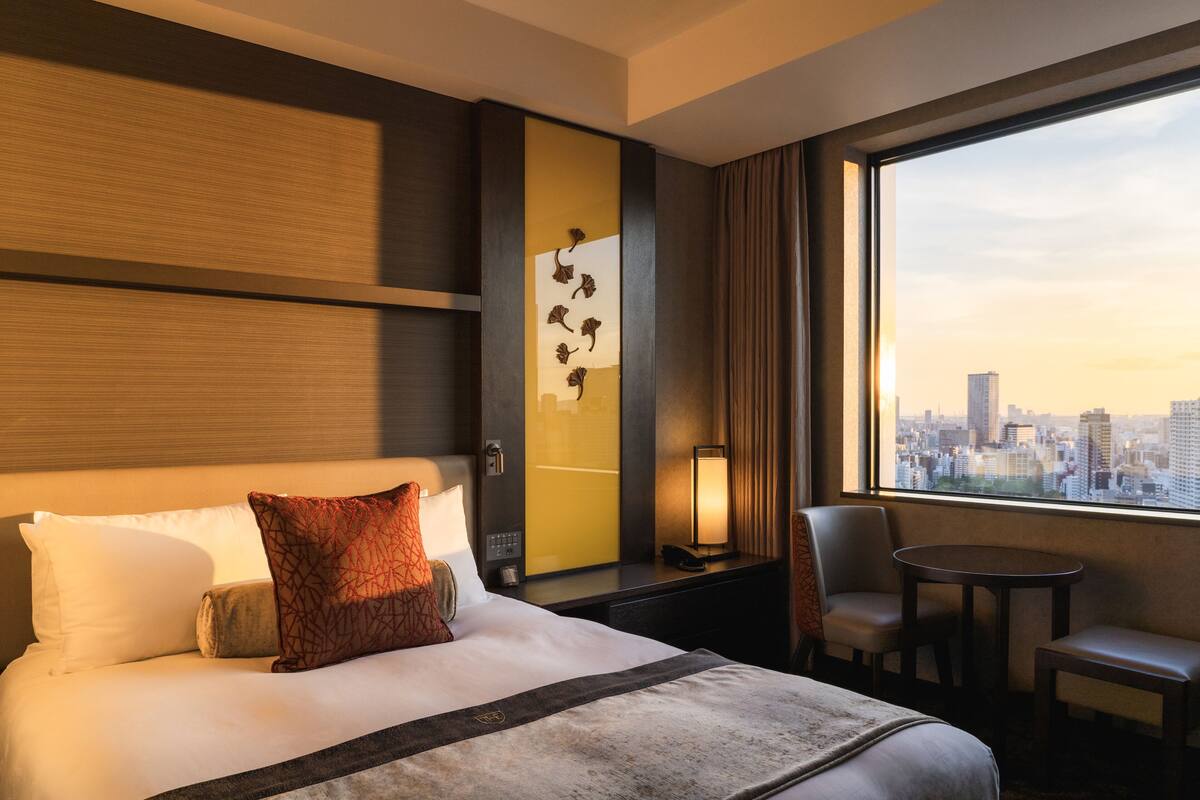A hotel room with a bed, a desk, a chair, and a view of a cityscape.