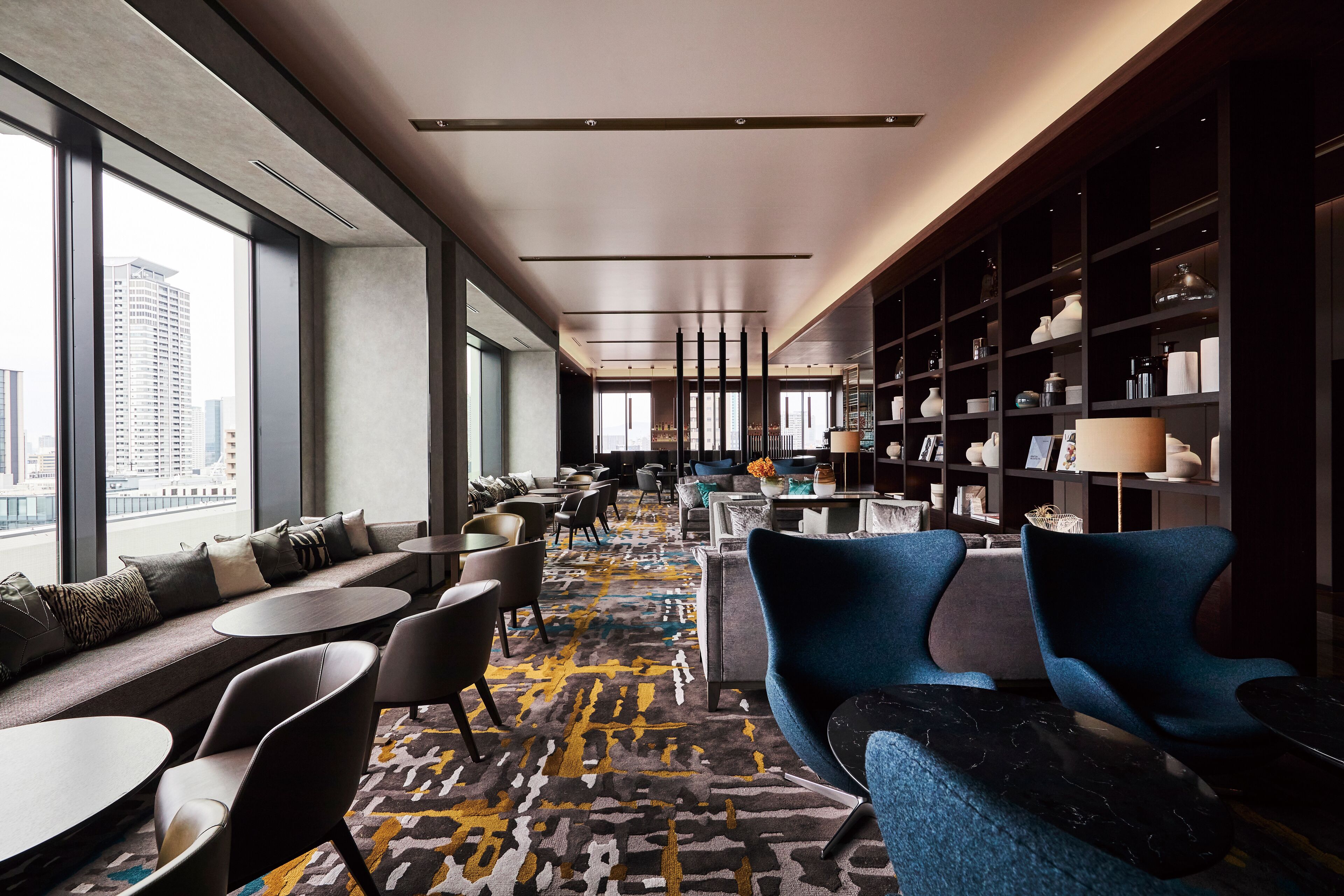 A modern hotel lobby with a long seating area, a bookshelf, and a view of the city.