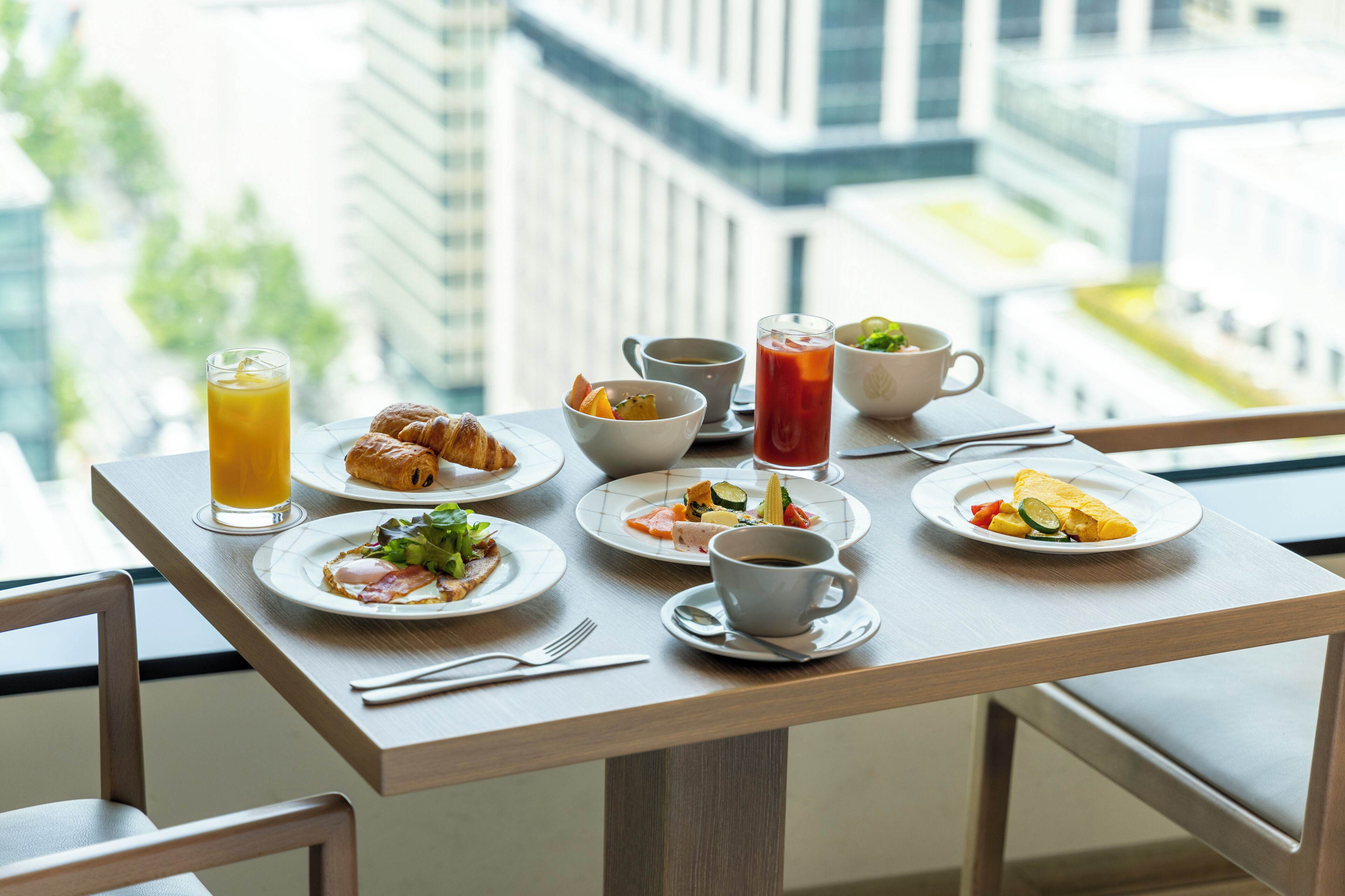 A breakfast table with various dishes and drinks, set against a city view.