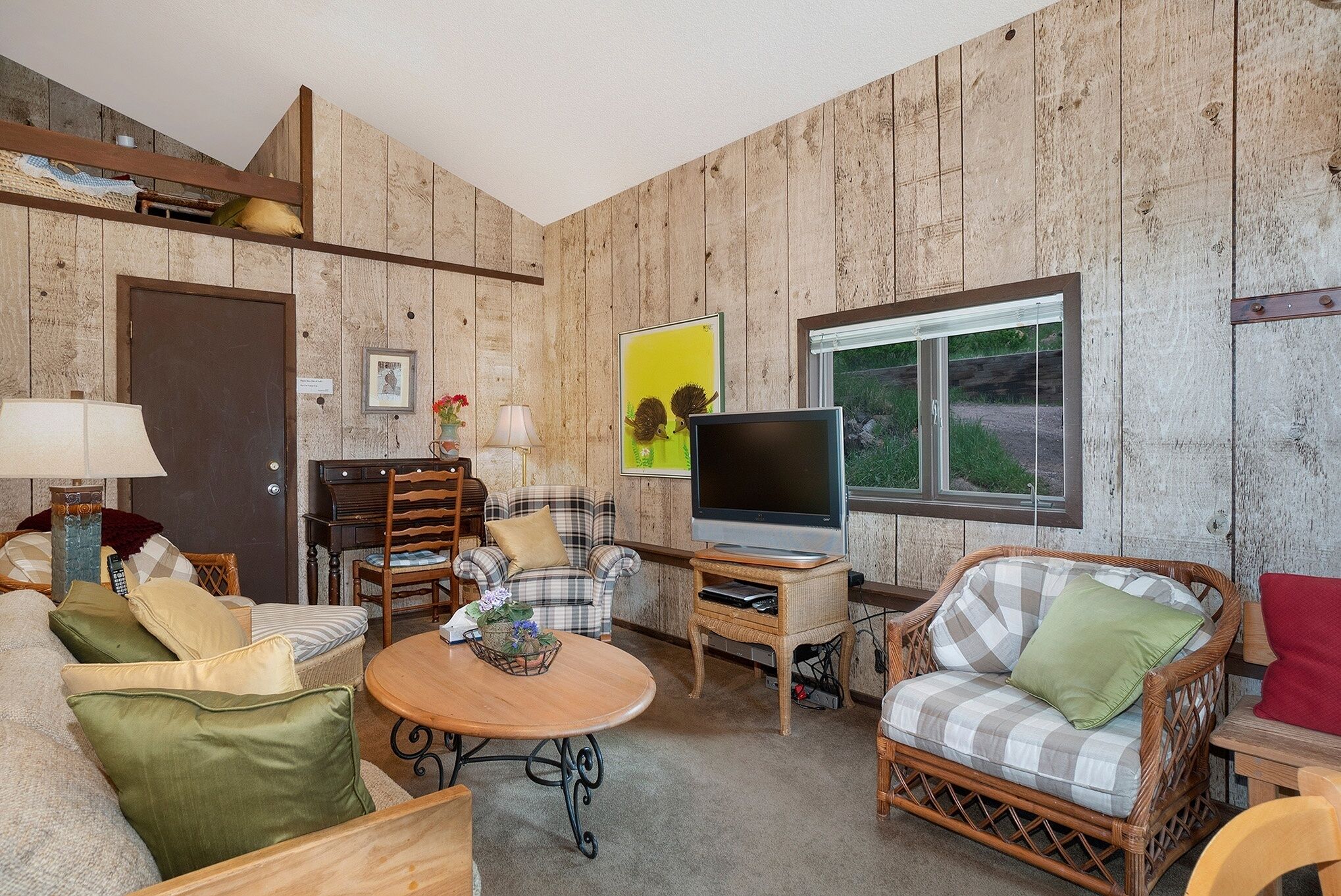 Un salon chaleureux avec des murs en bois, un téléviseur, des meubles en osier et une fenêtre donnant sur un beau paysage.