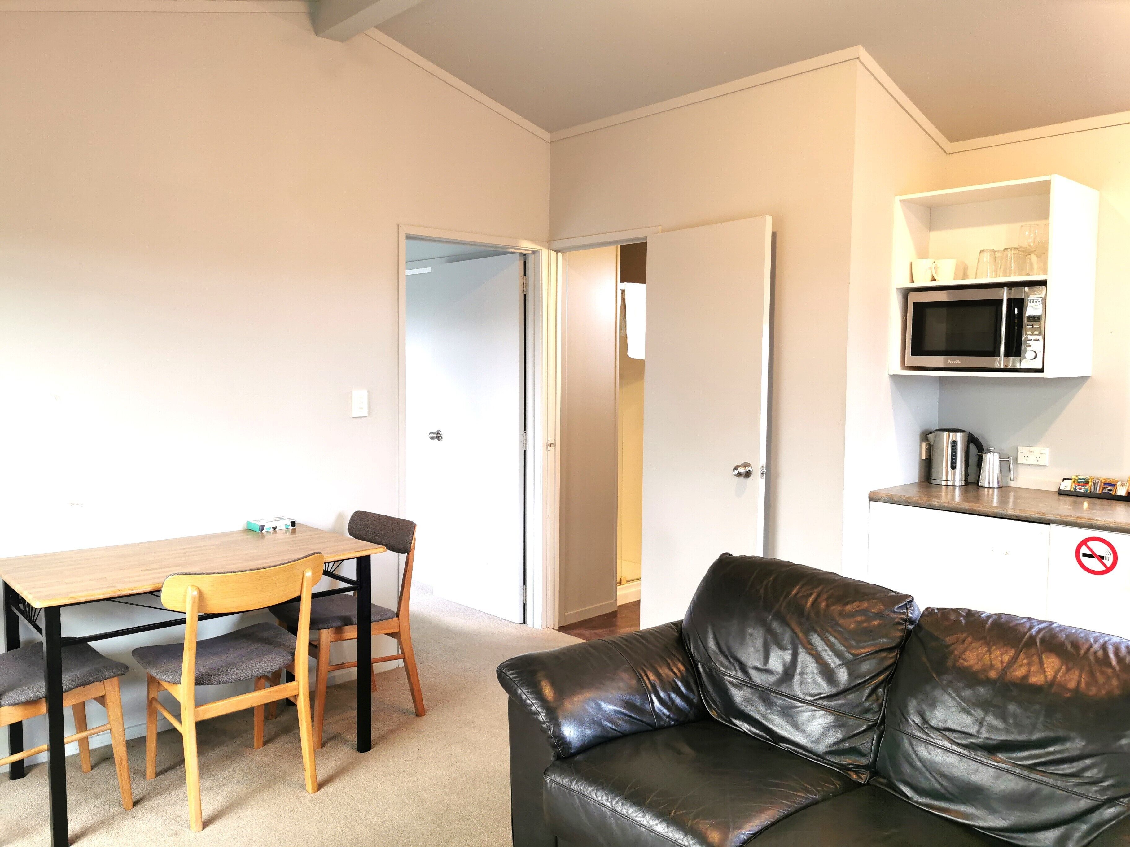 A small living area with a wooden dining table, two chairs, a black leather sofa, a kitchen area with a microwave, and a coffee maker.