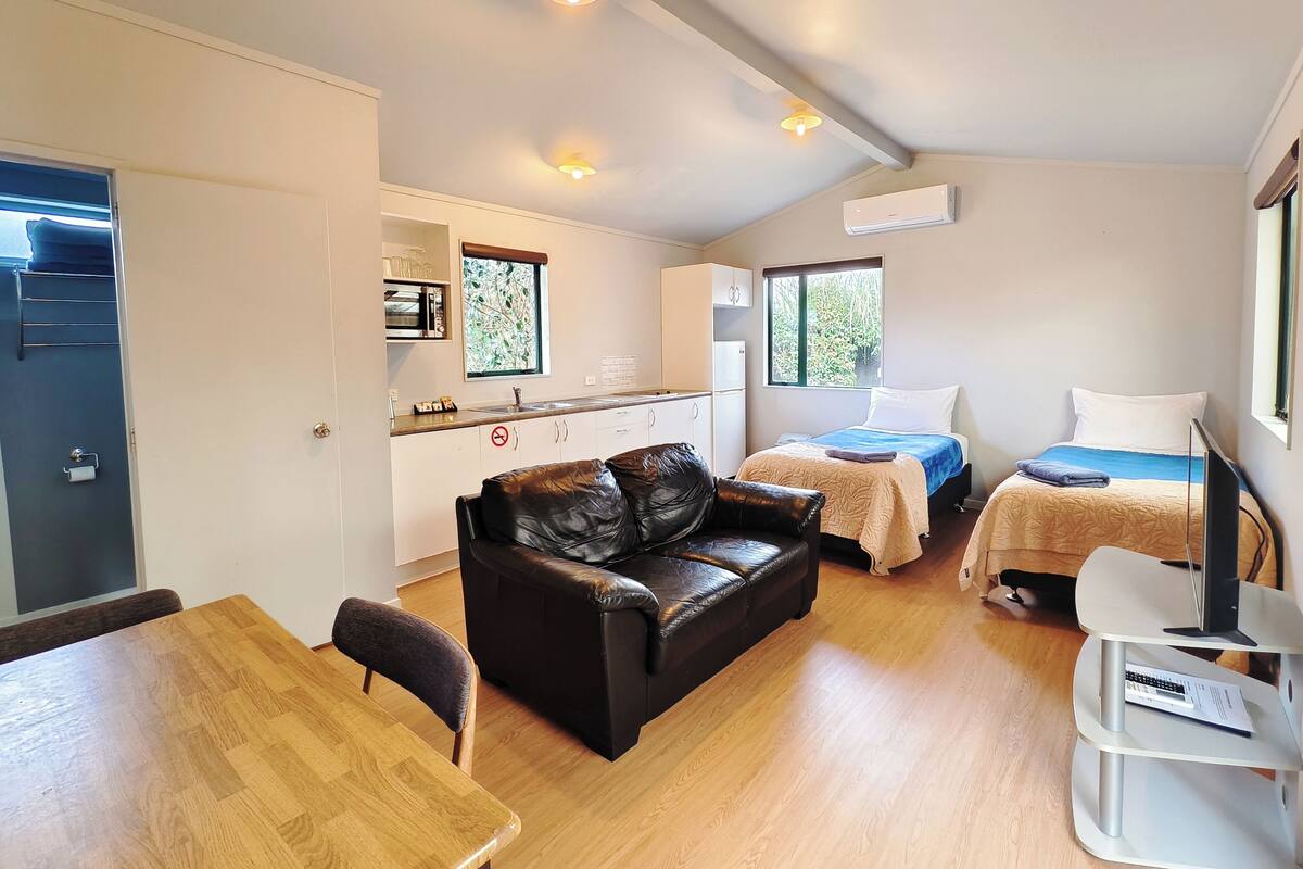 Una habitación compacta con kitchenette, una cama con cubrecama azul, un sofá de cuero, una mesa de comedor y un televisor sobre un pedestal.