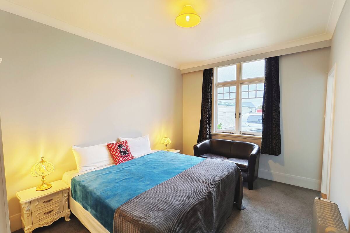 Una habitación de hotel con cama, mesitas de noche, una silla, ventana con cortinas y radiador.