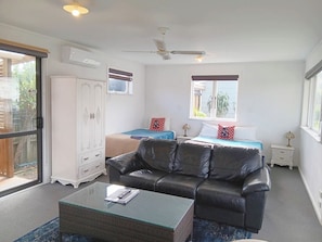 A room with a glass coffee table, a black leather sofa, two beds with patterned bedding, a white wardrobe, and a ceiling fan. - Parakai Geothermal Motel (Helensville)