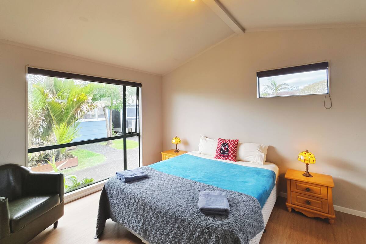 Un dormitorio con cama, escritorio, silla, ventana y una mesita de noche con lámpara.