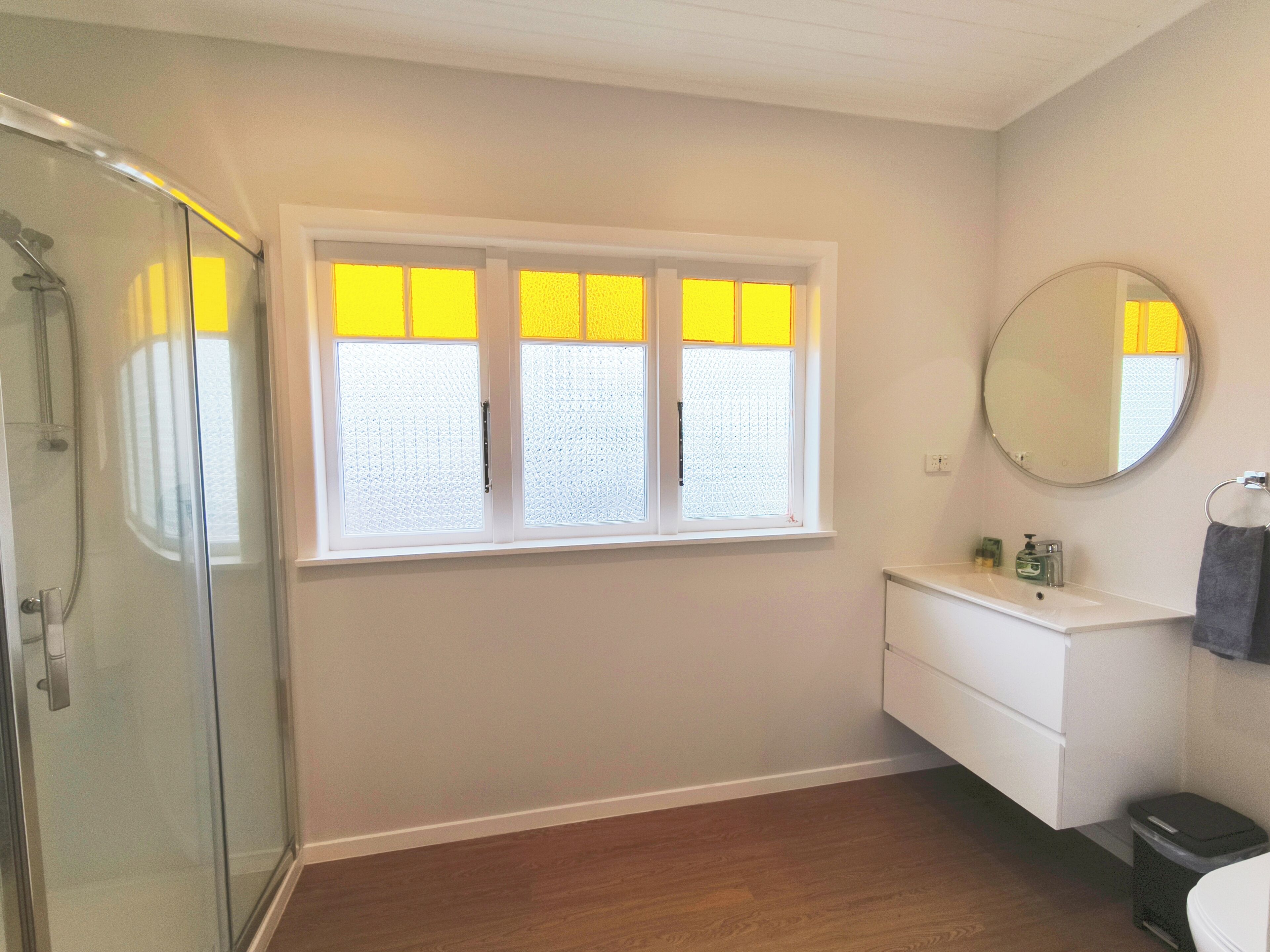 A bathroom with a glass shower enclosure, a white vanity with a mirror, and a window with yellow-tinted glass panes.