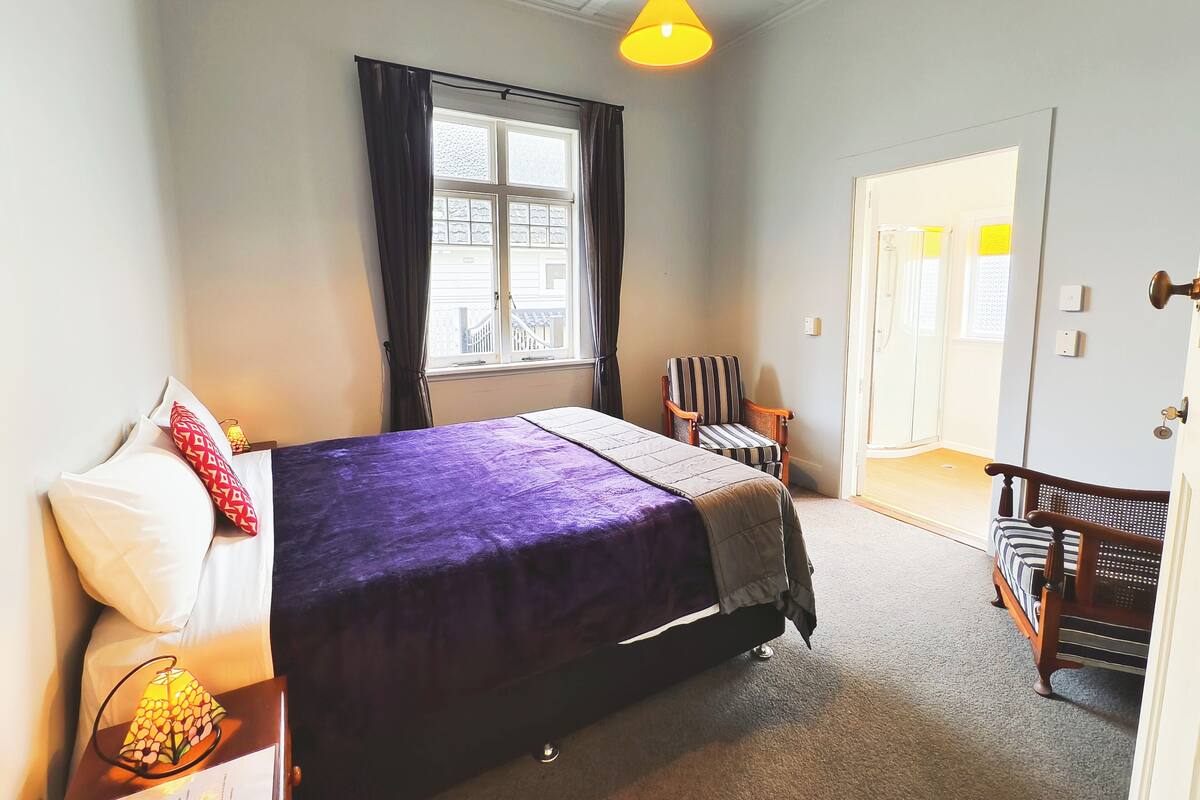 Un dormitorio con cama, una silla, una ventana con cortinas, una puerta y una lámpara.