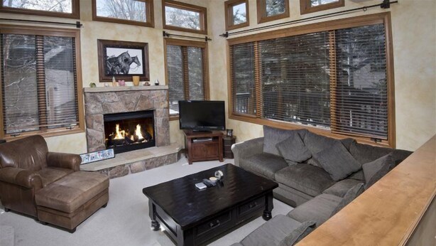 A living room with a stone fireplace, a television, and a large window with blinds. - Millers Creek Resident (Edwards)
