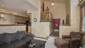 A spacious living room with a grey sofa, a brown leather chair, and a coffee table. A wooden staircase leads to a bedroom with a red bed. - Millers Creek Resident (Edwards)