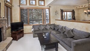 A living room with a sectional sofa, a flat-screen TV, a fireplace, and large windows with blinds. - Millers Creek Resident (Edwards)