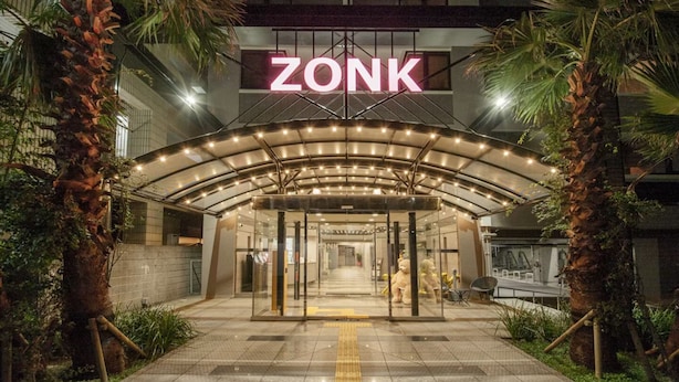 The entrance to a hotel named ZONK, featuring a glass facade, palm trees, and a covered entrance area. - Zonk Hotel Nakasu (Fukuoka)