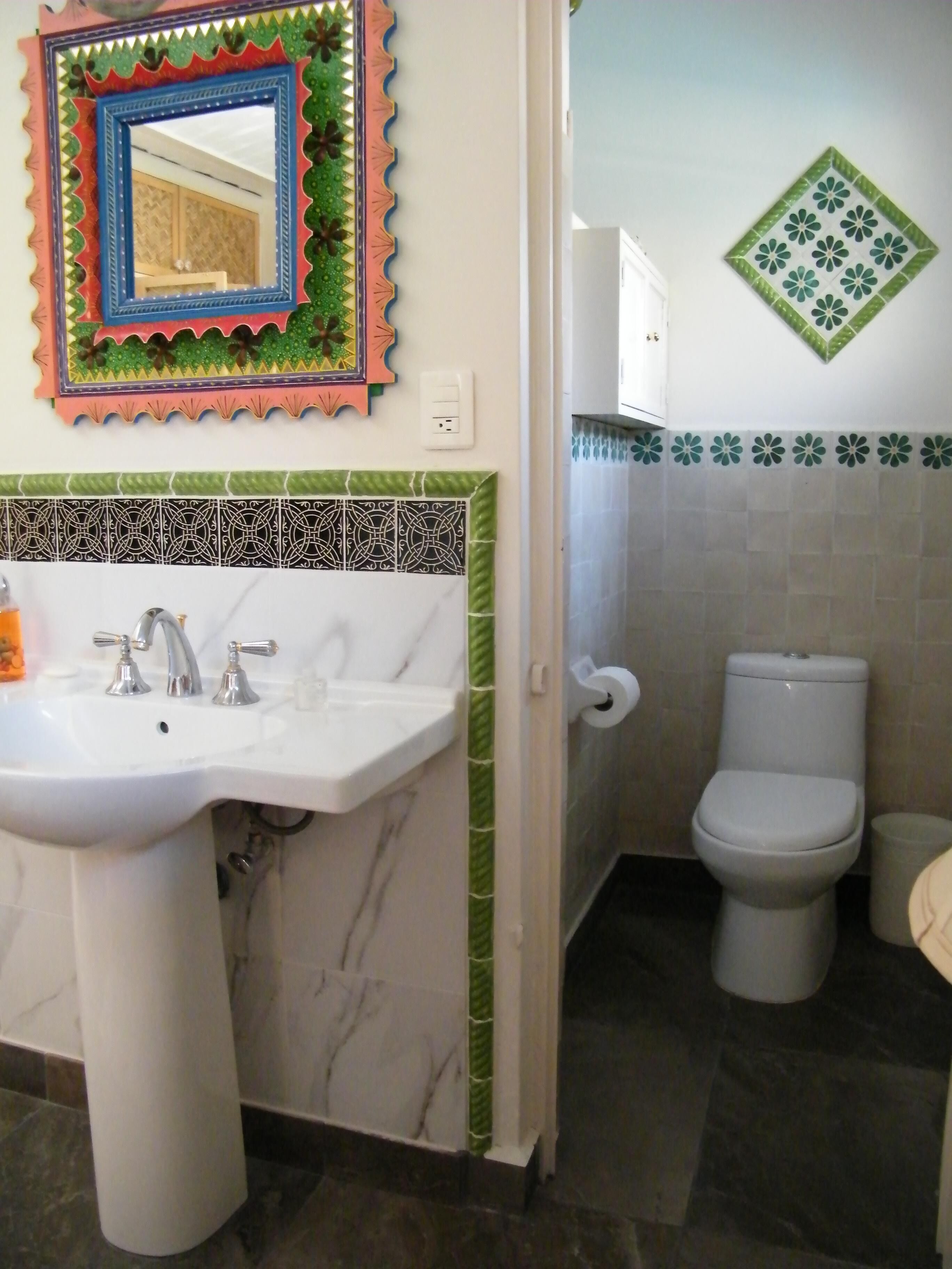 A bathroom with a white sink, two faucets, a mirror with a colorful frame, a toilet, and a trash bin.