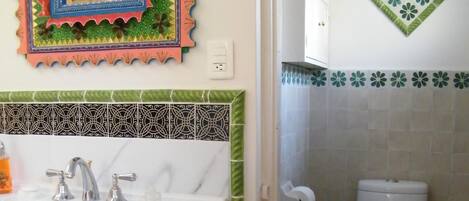 A bathroom with a white sink, two faucets, a mirror with a colorful frame, a toilet, and a trash bin.