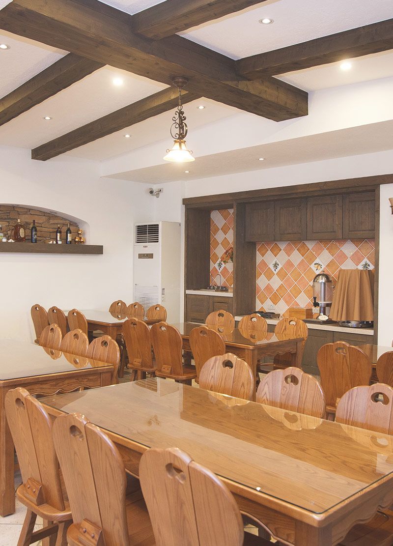 A dining area with a large wooden table and chairs, a hanging light fixture, and a kitchen in the background.