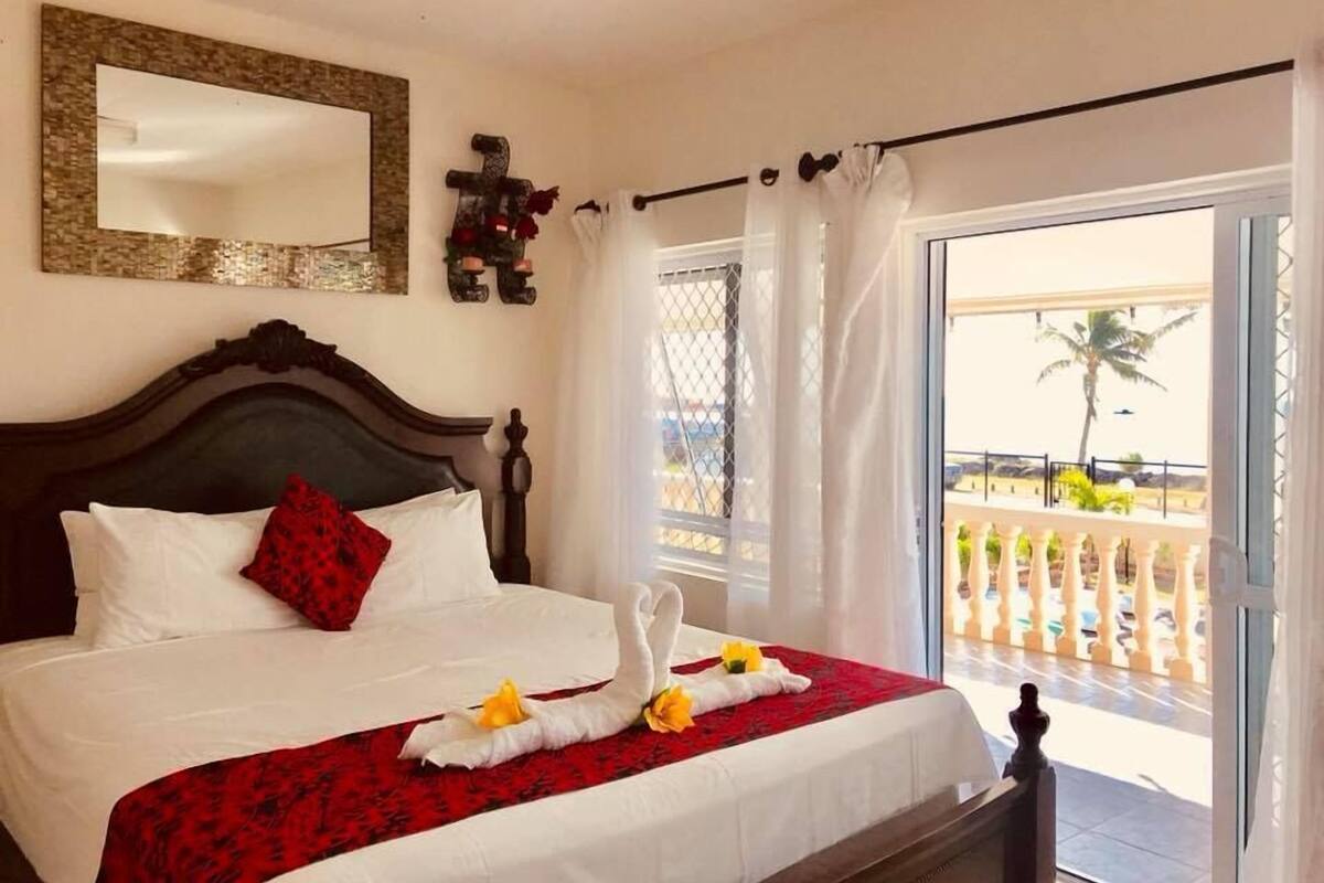 A hotel room with a large bed, a balcony with a view of palm trees, and a decorative cross on the wall.