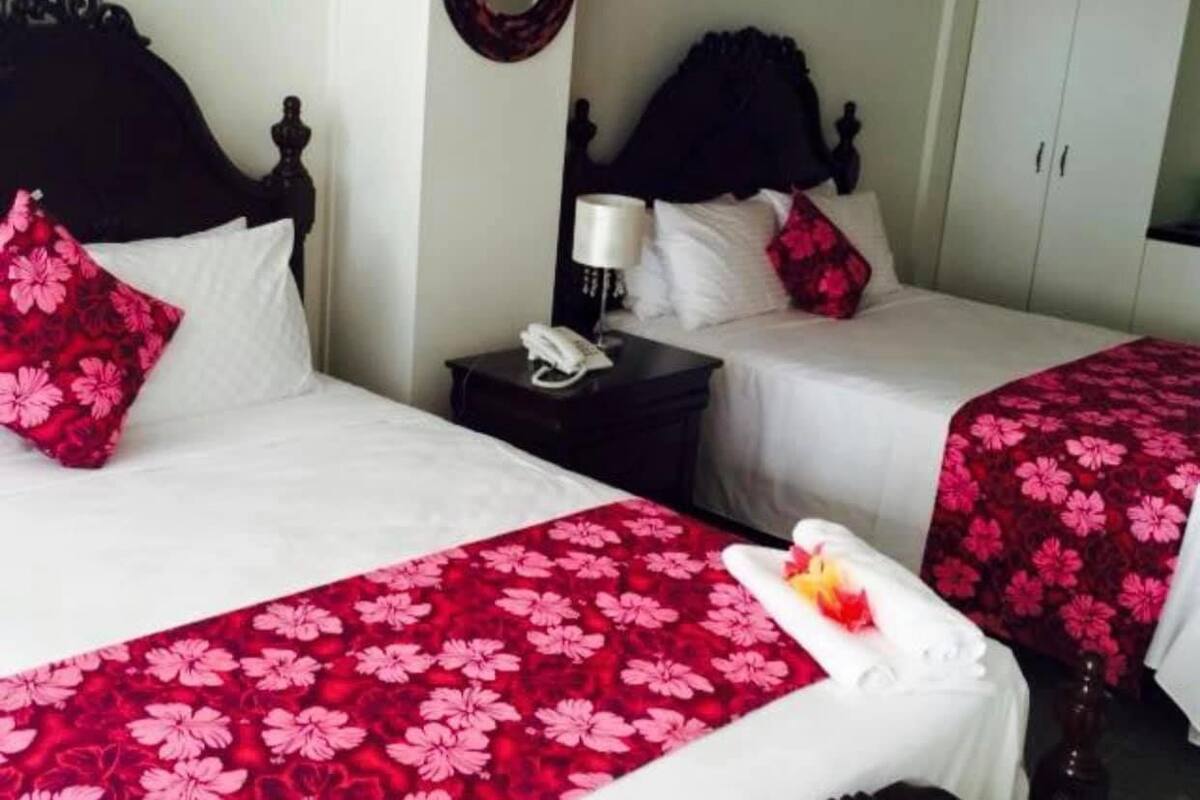 A hotel room with two beds, each with a red floral bedspread, white pillows, and a dark wooden headboard. There is a bedside table with a lamp and a phone.