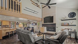 Un vaste espace de vie comprenant un grand canapé gris, une table basse noire et un ventilateur de plafond. On peut voir des paires de skis accrochées au mur et une cheminée en dessous.